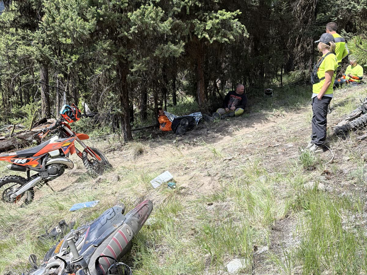 Medics in high-visibility gear attending to a rider resting in the trees beside a forest trail.