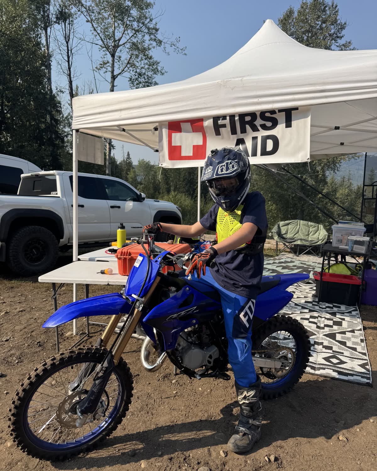 A young rider on a dirt bike stopped in front of the first aid canopy at an off-road event.